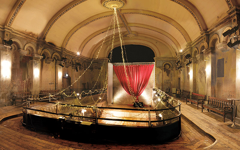 An ornate, historic hall with arched ceilings and worn walls, featuring benches along the sides and a central installation - airbnb guest tips london