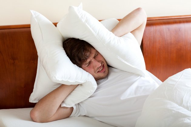 A man lying in bed covers his ears with pillows, appearing frustrated or disturbed, possibly due to loud noise or difficulty sleeping - airbnb neighbours