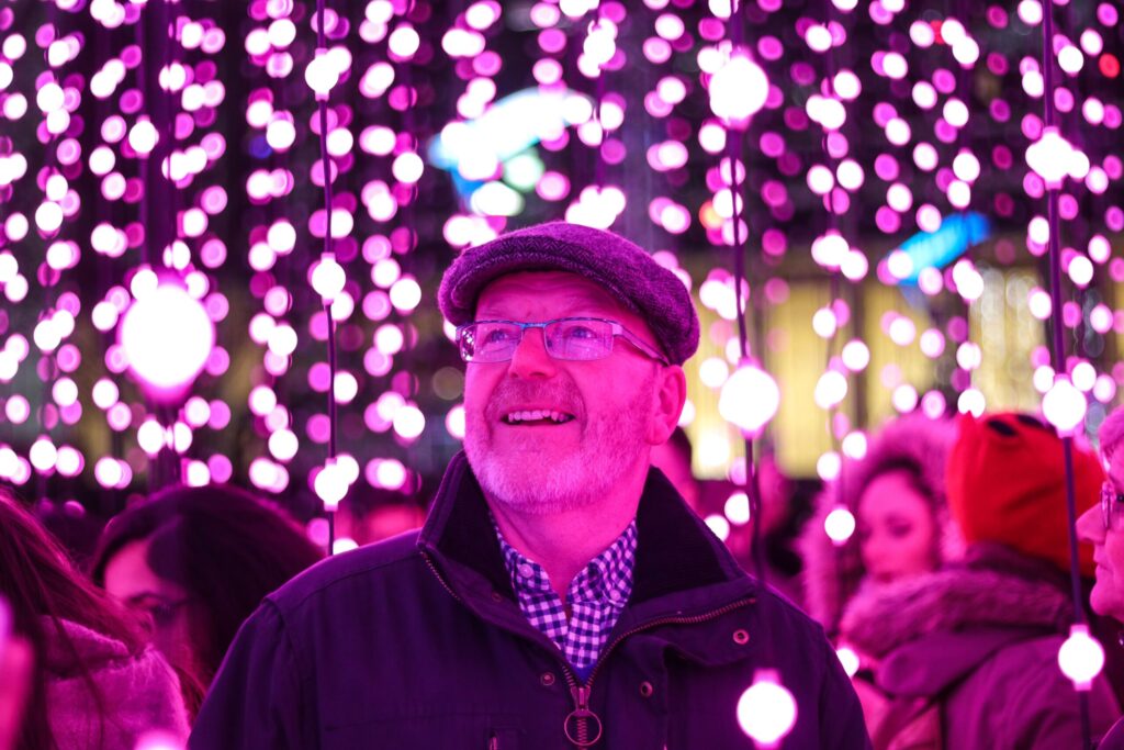 Un homme portant des lunettes, une casquette plate et une veste sourit, debout parmi des guirlandes lumineuses roses, entouré de gens dans un environnement vibrant et lumineux - hote airbnb senior