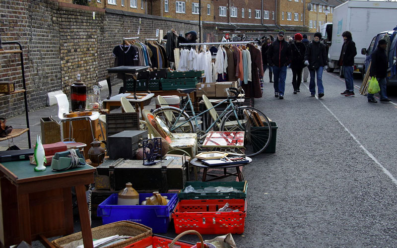 A street market with tables and crates displaying various secondhand items, including a bicycle, dishes, and clothing racks - airbnb guest tips london